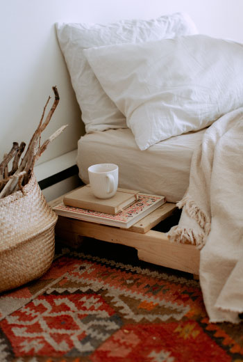 Un coin de lit blanc avec une petit table de nuit en blanc. Une tasse et des livres sont posés sur la table. Au sol un tapis boheme dans les tons rouges